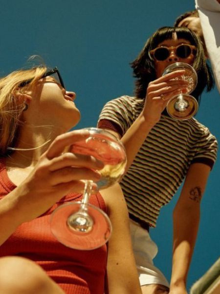 Two people outdoors under a clear blue sky holding stemmed glasses of spritz.