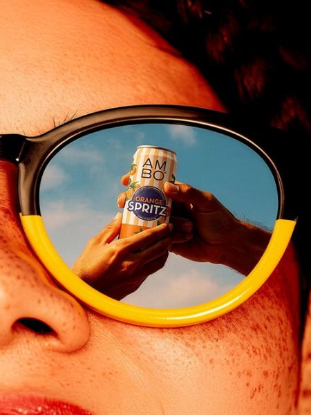 Hands holding an AMBO Spritz can reflected in yellow sunglasses against a blue sky.